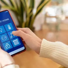 Mano usando un smartphone para controlar productos inteligentes de hogar: luces, temperatura, cámara, música y persianas.