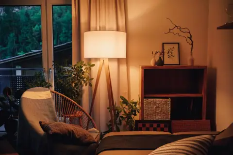 Lámpara de pie con luz blanco cálido ilumina un sillón acogedor y plantas. Estantería de madera con objetos decorativos y cestas.