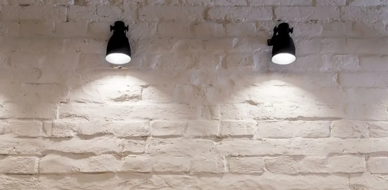 Dos lámparas de pared negras iluminan una pared de ladrillos blancos con un estilo rústico.