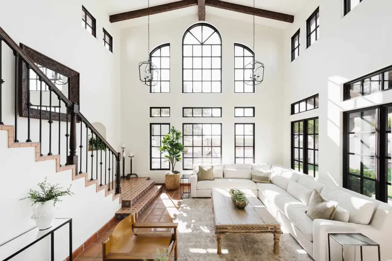 Amplio salón con techos altos, vigas de madera y grandes ventanales que inundan el espacio con luz neutra. Sofá blanco, mesa de centro rústica y escaleras con azulejos.